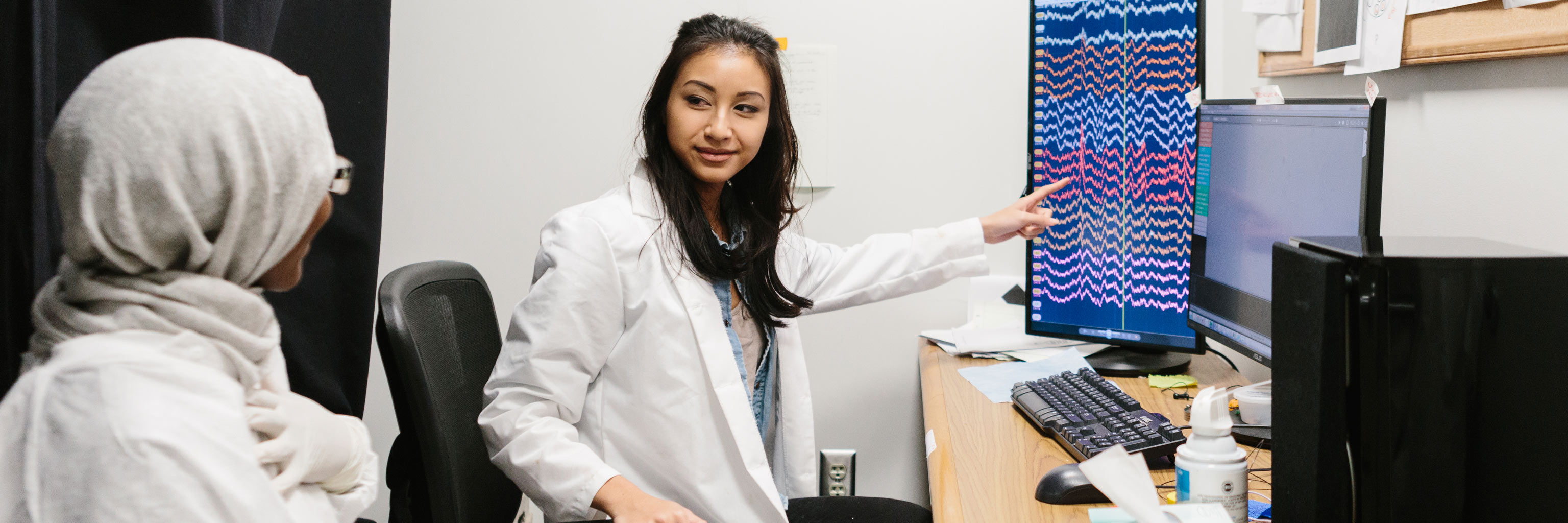 woman in white lab coat pointing at a computer screen talking to a cloaked woman